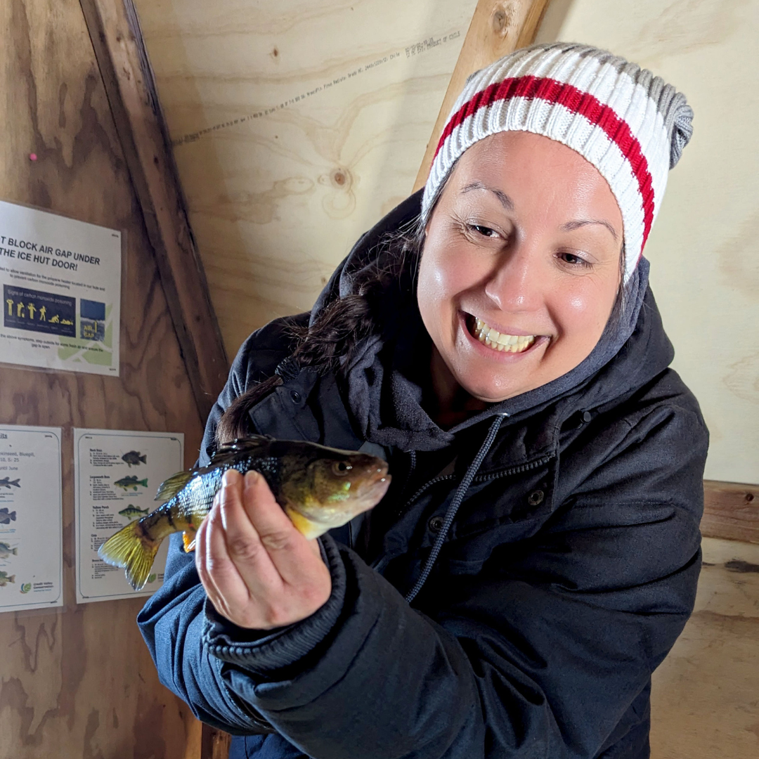 A woman dresses in winter apparel, smiles as she holds a fish while sitting in a fish hut.